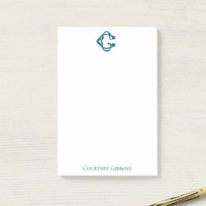 May include: A white notepad with a blue monogram of the letter "C" and the name "COURTNEY GIBBONS" printed at the bottom. A yellow pencil is lying on a white wooden surface.