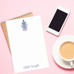 May include: A white notepad with a blue and white patterned porcelain jar design and the name "Beth Wright" printed at the bottom. The notepad is on a pink surface with a brown notepad, a white smartphone, and a white saucer with a cup of coffee.