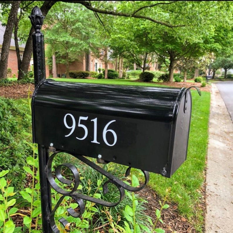 May include: Black mailbox with the number 9516 in white lettering. The mailbox is mounted on a black metal post with decorative scrollwork. The background includes green foliage, trees, and a residential street.