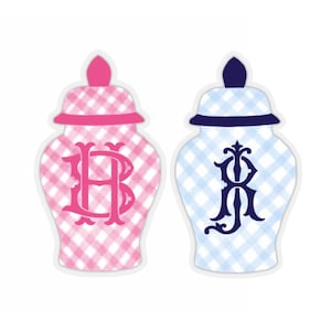 May include: Two decorative ceramic ginger jars with a gingham pattern. The jar on the left is pink with a white gingham pattern and a pink lid. The jar on the right is blue with a white gingham pattern and a blue lid. Both jars have a monogrammed initial on the front.
