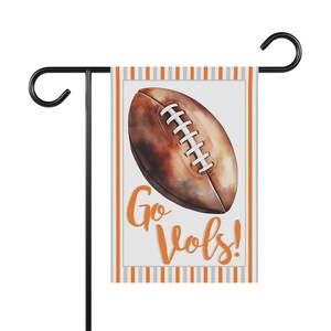 Puede incluir: Una bandera de jard&iacute;n con un bal&oacute;n de f&uacute;tbol de estilo acuarela en tonos marr&oacute;n y blanco. La bandera tiene un fondo blanco con las palabras "Go Vols!" en escritura naranja. La bandera est&aacute; bordeada por rayas naranjas y grises.