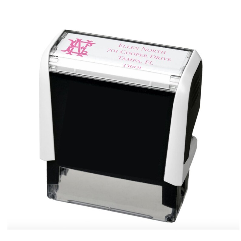 K&ouml;nnte beinhalten: Ein selbstf&auml;rbender Adressstempel mit wei&szlig;em und schwarzem Geh&auml;use. Die Oberseite zeigt ein rosa Monogramm und Adressdetails: Ellen North, 701 Cooper Drive, Tampa, FL 33601. Der Stempel ist f&uuml;r einen klaren und effizienten Abdruck konzipiert.