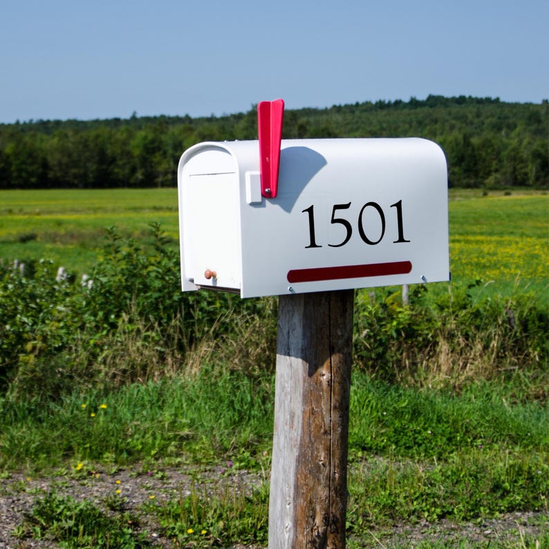 May include: White mailbox with the number 1501 in black lettering. The mailbox has a red flag and is mounted on a wooden post. The background features green fields and trees under a blue sky.