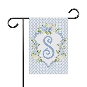 May include: A blue and white garden flag with a white shield featuring a blue letter "S" surrounded by blue and white flowers. The background is a blue and white checkered pattern.