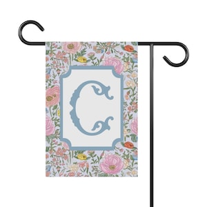 May include: A garden flag with a floral pattern in shades of pink, yellow, and green. The flag features a blue letter C in a white frame.