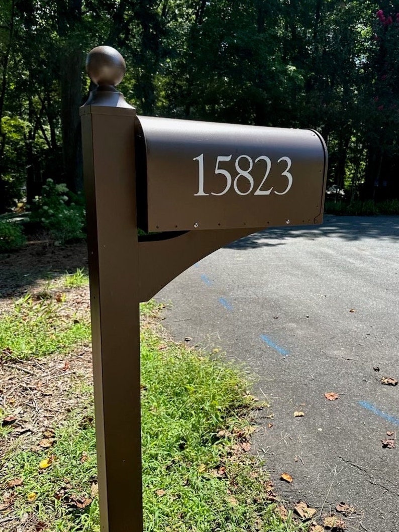 May include: A brown metal mailbox with the number "15823" in white lettering. The mailbox has a rounded top and a decorative finial. It is mounted on a matching brown post and is surrounded by greenery.