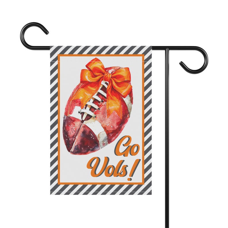 Puede incluir: Una bandera de jard&iacute;n con un dise&ntilde;o de f&uacute;tbol en acuarela, un lazo naranja y las palabras "Go Vols!". La bandera tiene un borde a rayas blanco y negro y un marco naranja. Est&aacute; sujeta a un soporte de metal negro.