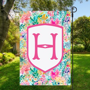 Peut inclure: Un drapeau de jardin rose et blanc avec un motif floral et une grande lettre rose "H" au centre. Le drapeau est suspendu &agrave; un poteau en m&eacute;tal noir.