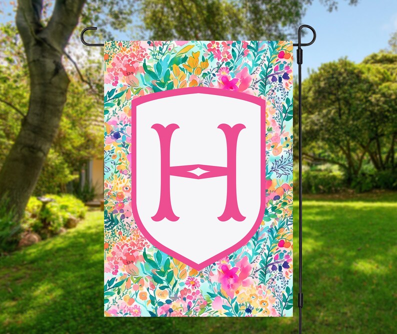 Peut inclure: Un drapeau de jardin rose et blanc avec un motif floral et une grande lettre rose "H" au centre. Le drapeau est suspendu &agrave; un poteau en m&eacute;tal noir.