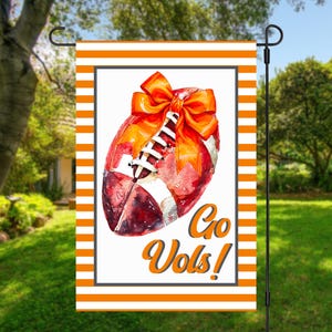 Puede incluir: Una bandera de jard&iacute;n con un dise&ntilde;o de f&uacute;tbol en acuarela, un lazo naranja y las palabras "Go Vols!" en escritura naranja. La bandera tiene un borde a rayas naranja y blanco y se muestra en un soporte de metal negro.