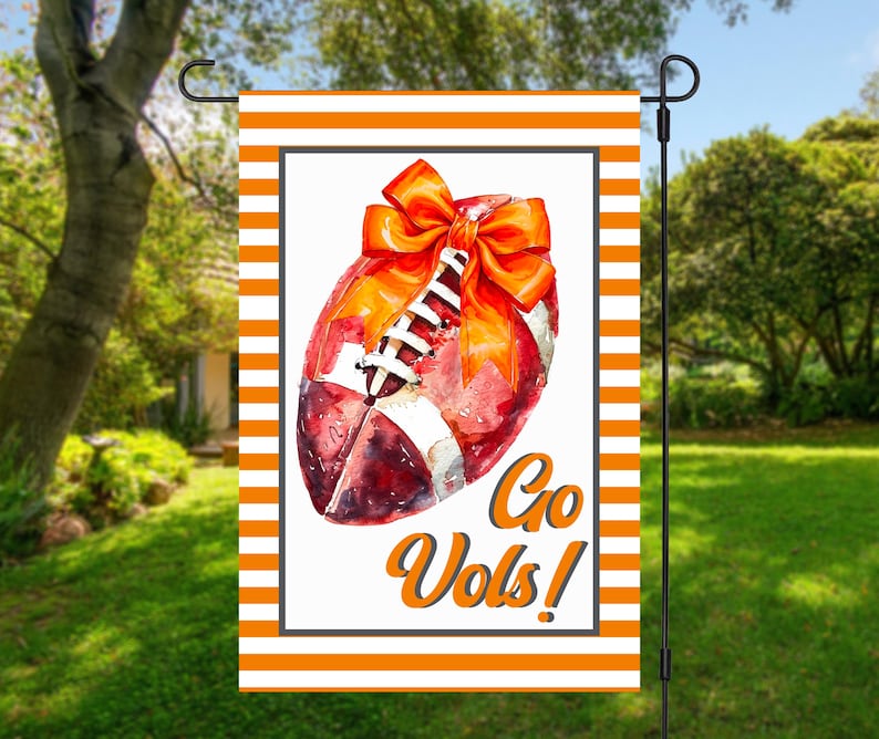 Puede incluir: Una bandera de jard&iacute;n con un dise&ntilde;o de f&uacute;tbol en acuarela, un lazo naranja y las palabras "Go Vols!" en escritura naranja. La bandera tiene un borde a rayas naranja y blanco y se muestra en un soporte de metal negro.