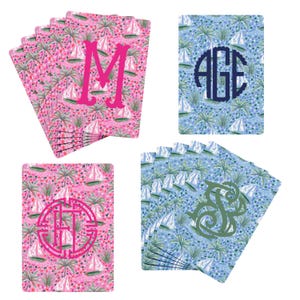 May include: A collection of rectangular playing cards featuring a repeating pattern of sailboats and palm trees. Some cards are pink with a hot pink letter M or a circular monogram, while others are blue with a dark blue monogram or green script.