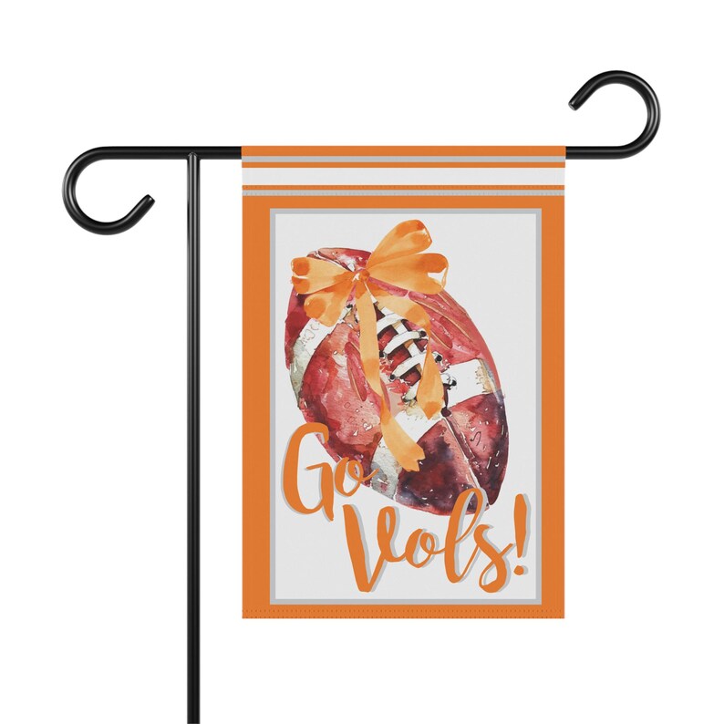 Puede incluir: Una bandera de jard&iacute;n con un bal&oacute;n de f&uacute;tbol de estilo acuarela, un lazo naranja y las palabras "Go Vols!" en escritura naranja. La bandera tiene un borde naranja y est&aacute; montada en un soporte de metal negro.