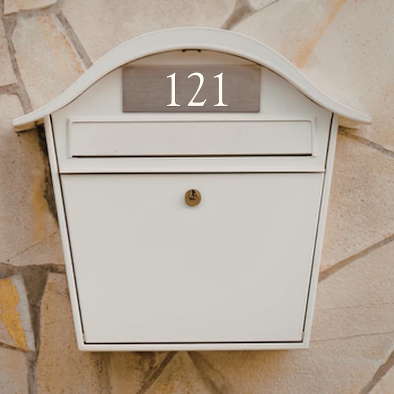 May include: A white mailbox with the number 121 displayed on a small, rectangular plaque. The mailbox has a curved top and a small lock. The background is a stone wall.