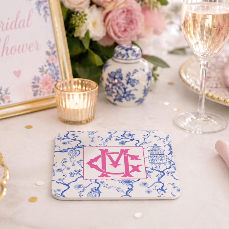 May include: A square white coaster with a blue floral pattern and a pink monogram. A lit candle in a gold votive, a blue and white jar, and a glass of champagne are on a white tablecloth. A sign that says "bridal shower" is in the background.