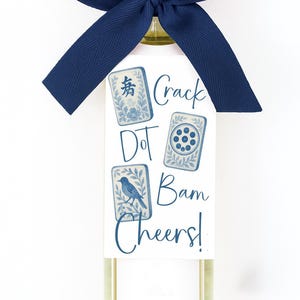 May include: A bottle of white wine with a navy blue bow and a tag featuring Mahjong tile illustrations. The tag reads "Crack Dot Bam Cheers!" in navy blue script. The bottle has a silver cap.