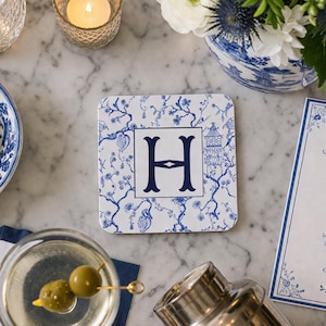 May include: Square white coaster with a blue floral pattern and a large navy blue letter "H" in the center. The coaster is surrounded by a martini, flowers, and a lit candle on a marble surface.