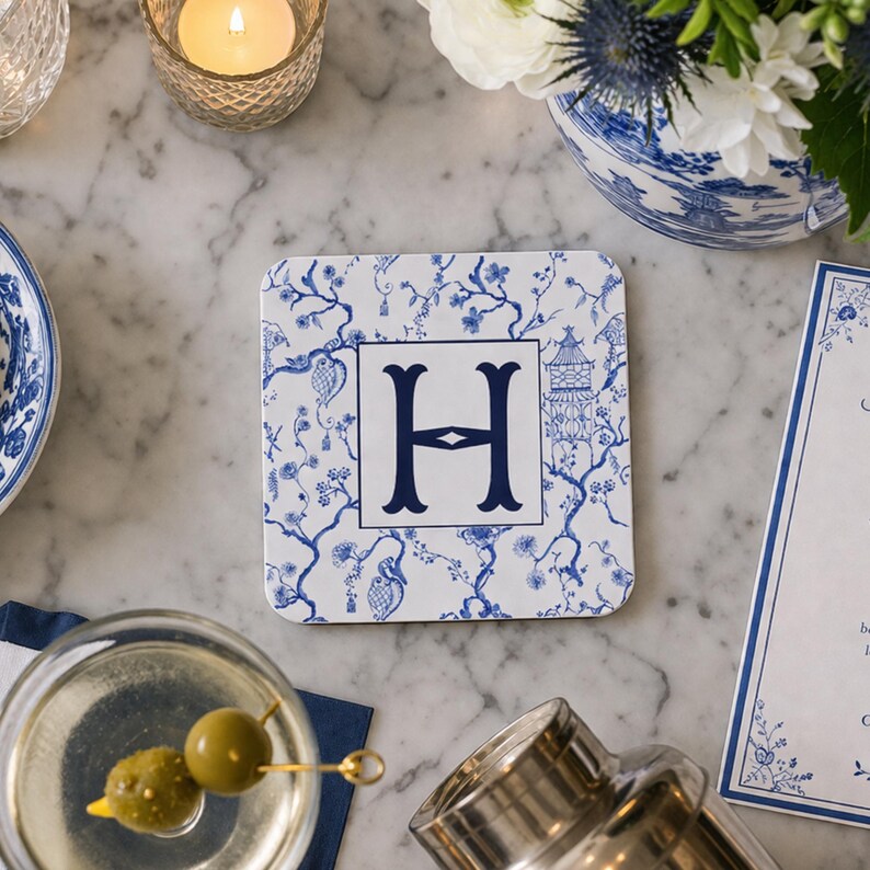 May include: Square white coaster with a blue floral pattern and a large navy blue letter "H" in the center. The coaster is surrounded by a martini, flowers, and a lit candle on a marble surface.