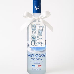 May include: A frosted Grey Goose Vodka bottle with a blue cap and a white ribbon tied around the neck. A tag with the words "Crack Dot Bam Cheers!" is attached. The bottle has the words "Grey Goose Vodka" printed on it, and the French flag is at the bottom.