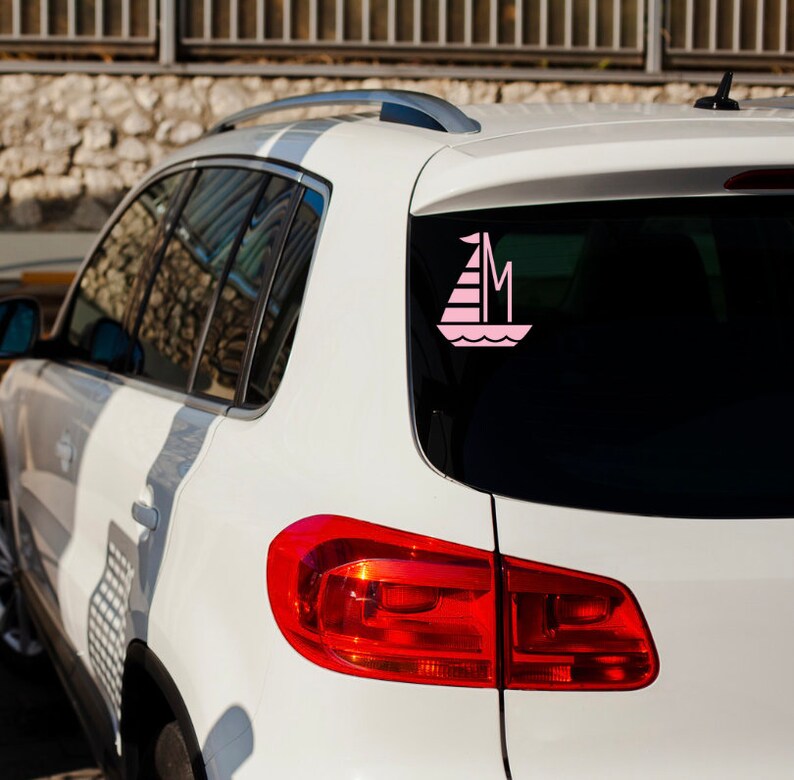 K&ouml;nnte beinhalten: Ein wei&szlig;es Auto mit einem rosa Segelboot-Aufkleber auf der Heckscheibe. Der Aufkleber hat den Buchstaben "M" auf dem Segel.