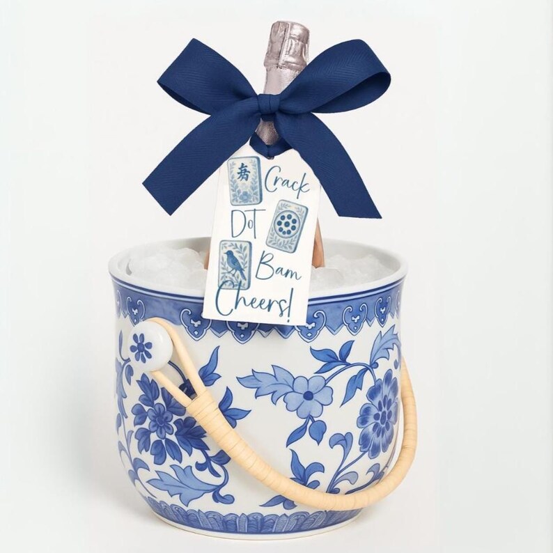 May include: A white and blue floral patterned ice bucket with a woven handle holds ice and a champagne bottle. A navy blue bow is tied around the bottle's neck, with a tag that reads "Crack Dot Bam Cheers!"