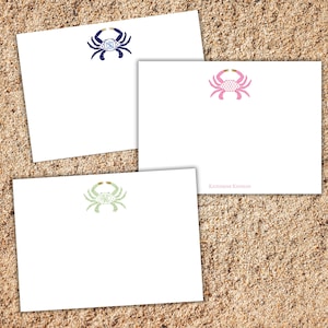 May include: Three white stationery cards with crab designs in navy blue, pink, and green. Each crab has a gold-colored accent and a monogram. The cards are arranged on a sandy surface.