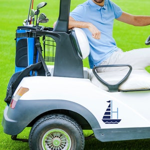 K&ouml;nnte beinhalten: Ein wei&szlig;er Golfwagen mit einem blauen und wei&szlig;en Segelboot-Aufkleber an der Seite. Der Wagen hat eine schwarze Golftasche mit blauen Akzenten im R&uuml;cken. Eine Person sitzt im Wagen.