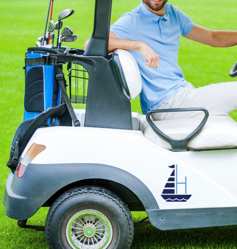 K&ouml;nnte beinhalten: Ein wei&szlig;er Golfwagen mit einem blauen und wei&szlig;en Segelboot-Aufkleber an der Seite. Der Wagen hat eine schwarze Golftasche mit blauen Akzenten im R&uuml;cken. Eine Person sitzt im Wagen.
