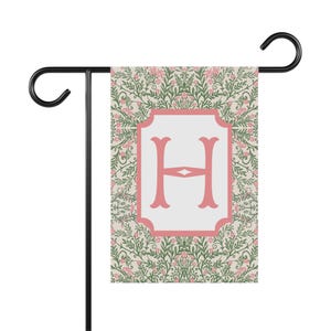 May include: A pink and green garden flag with a white monogram of the letter "H" in a pink frame. The flag has a floral pattern with pink flowers and green leaves.