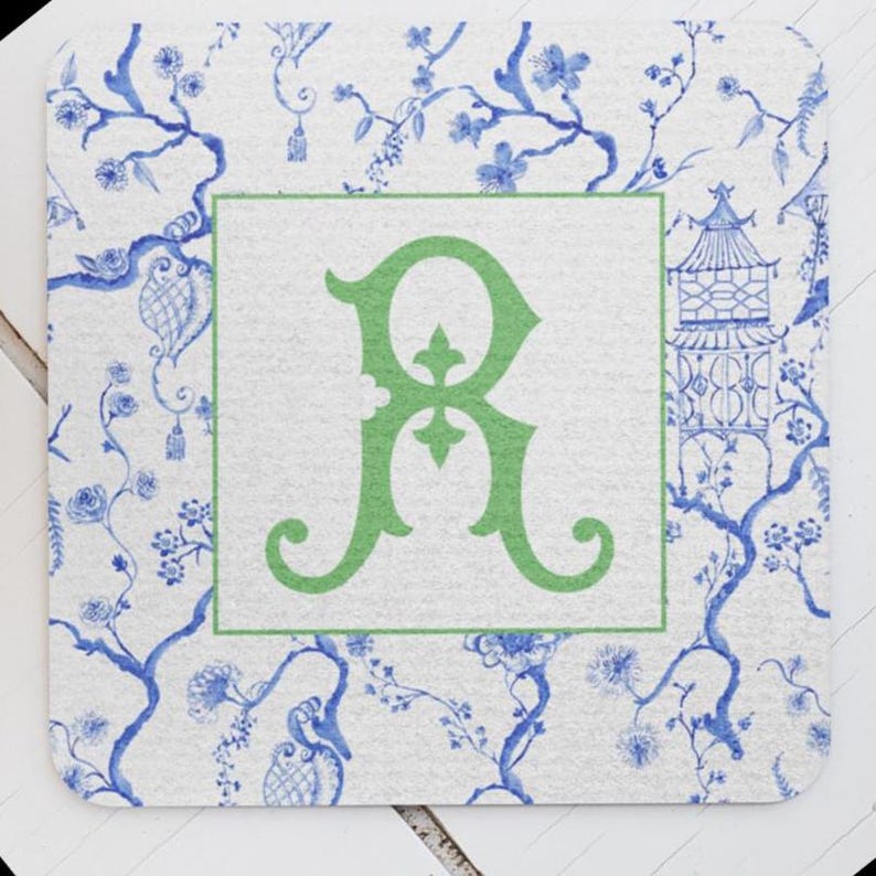 May include: Square coaster with a white background featuring a blue floral pattern and a green border. The center has a green letter "R". The coaster has a classic, elegant design.