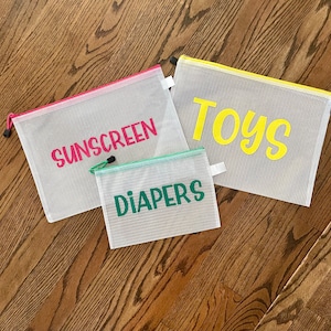 May include: Three white mesh zipper pouches with colorful trim and labels. The labels read "SUNSCREEN" in pink, "TOYS" in yellow, and "DIAPERS" in green. The pouches are arranged on a wooden surface.