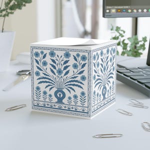 May include: A white square notepad with a blue floral design. The design features a vase with flowers and leaves, surrounded by a border of smaller flowers. The notepad is on a white desk with paperclips and a computer.