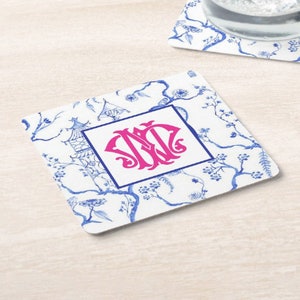 May include: Square coaster with a white background featuring a blue floral and pagoda design. A pink monogram is centered within a blue square. The coaster is likely made of ceramic or a similar material.