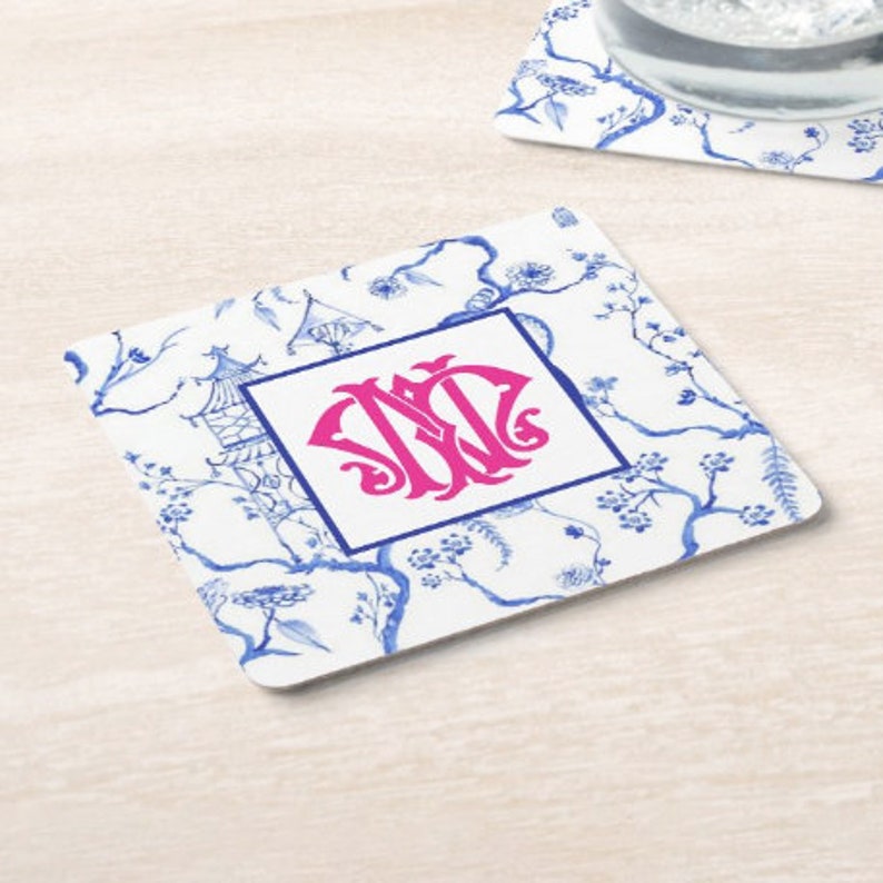 May include: Square coaster with a white background featuring a blue floral and pagoda design. A pink monogram is centered within a blue square. The coaster is likely made of ceramic or a similar material.