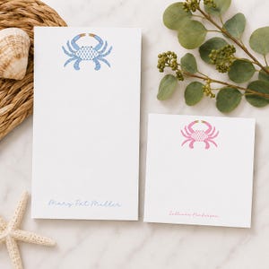May include: Two white notepads with crab designs. One notepad has a blue crab design and the name "Mary Pat Miller" in blue script. The other notepad has a pink crab design and the name "Sullivan Henderson" in pink script.