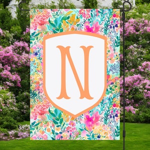 Peut inclure: Un drapeau de jardin avec un bouclier blanc sur lequel est inscrit la lettre "N" en couleur p&ecirc;che. Le bouclier est entour&eacute; d'un motif floral color&eacute; avec des nuances de rose, jaune, bleu et vert.