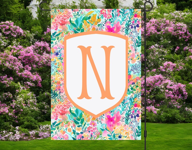 Peut inclure: Un drapeau de jardin avec un bouclier blanc sur lequel est inscrit la lettre "N" en couleur p&ecirc;che. Le bouclier est entour&eacute; d'un motif floral color&eacute; avec des nuances de rose, jaune, bleu et vert.
