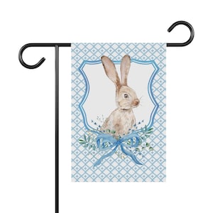 Peut inclure: Un drapeau de jardin bleu et blanc avec une illustration aquarelle d'un lapin brun avec un nœud bleu. Le lapin est entouré de feuilles vertes et de fleurs.