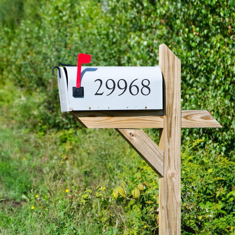 May include: A white mailbox with the number "29968" in black. The mailbox has a red flag and is mounted on a wooden post. The background is filled with green foliage.