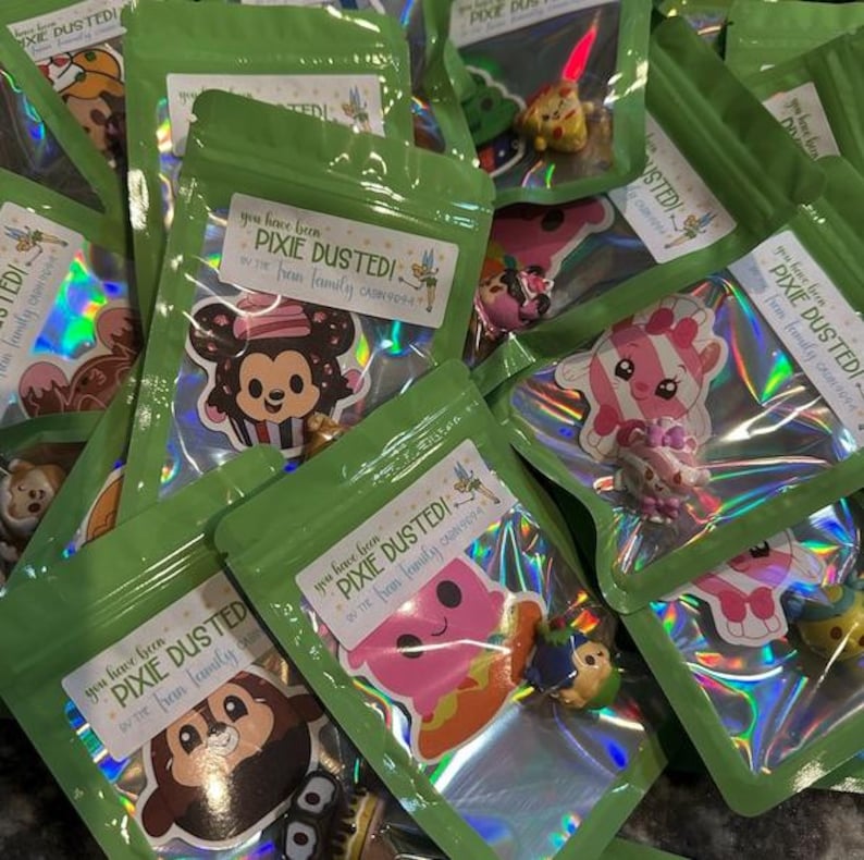 May include: A pile of small green bags with holographic iridescent lining. Each bag has a sticker with a cartoon character and the text "You have been Pixie Dusted! by the [family name] family".