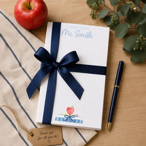 May include: A white notepad with a navy blue ribbon and bow, featuring the text "A Note From Ms. Smith." An apple and pen are next to the notepad. The notepad has a watercolor illustration of an apple and books. A tag reads "Thank you for all you do!"