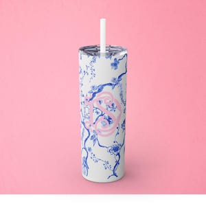 May include: A white stainless steel tumbler with a blue and white floral pattern and a pink monogram. The tumbler has a straw.