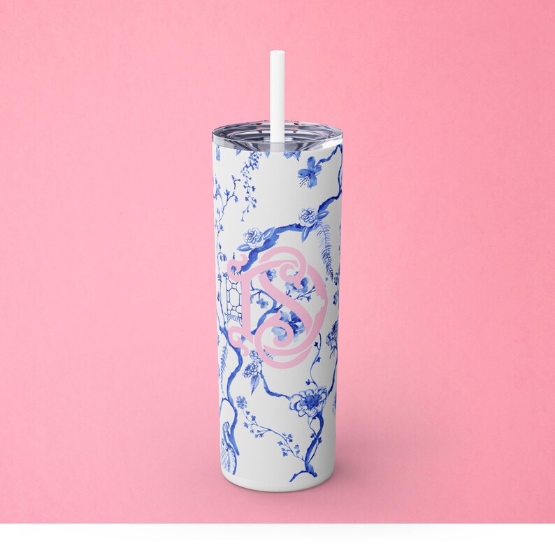 May include: A white stainless steel tumbler with a blue and white floral pattern and a pink monogram. The tumbler has a straw.