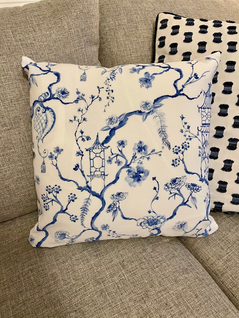 May include: A white throw pillow with a blue floral pattern featuring branches, flowers, and pagodas. The pillow is on a gray couch.