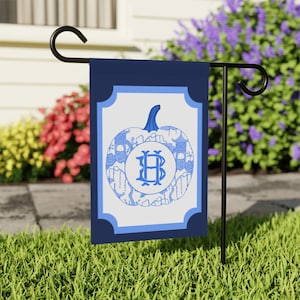May include: A blue and white garden flag with a blue pumpkin design and the monogram "HB" in the center. The flag is attached to a black metal garden flag stand.