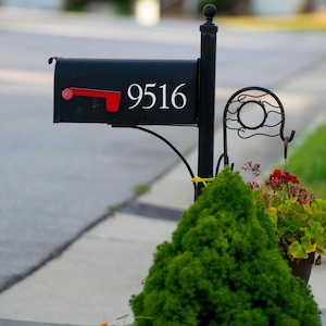 May include: A black mailbox with the number 9516 in white. The mailbox has a red flag and is mounted on a black post. A small green bush and flowers are in front of the mailbox.