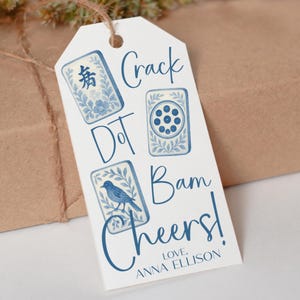 May include: A white gift tag with blue illustrations of game tiles and a bird, with the words "Crack Dot Bam Cheers!" and "Love, Anna Ellison" in blue script. The tag is tied with twine.