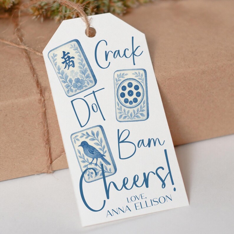 May include: A white gift tag with blue illustrations of game tiles and a bird, with the words "Crack Dot Bam Cheers!" and "Love, Anna Ellison" in blue script. The tag is tied with twine.