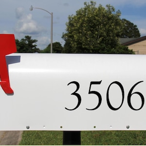 May include: A white mailbox with the number 3506 in black. The mailbox has a red flag on the side, indicating mail delivery. The scene is outdoors on a sunny day.