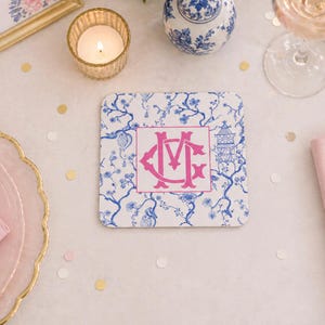 May include: Square white coaster with a blue floral pattern and a pink monogram. The coaster has a pagoda and floral design. A lit candle, a glass of wine, and decorative items are in the background.
